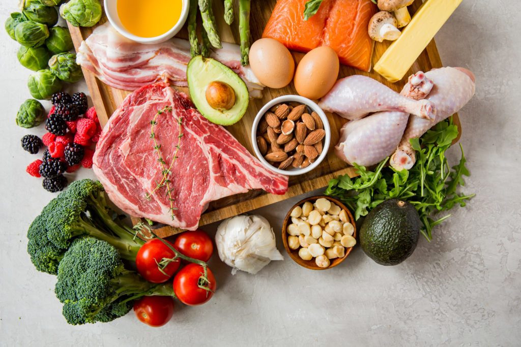 A spread of keto friendly foods including steak, nuts, avocado broccoli, berries, salmon, eggs, cheese, bacon, oils, and greens.