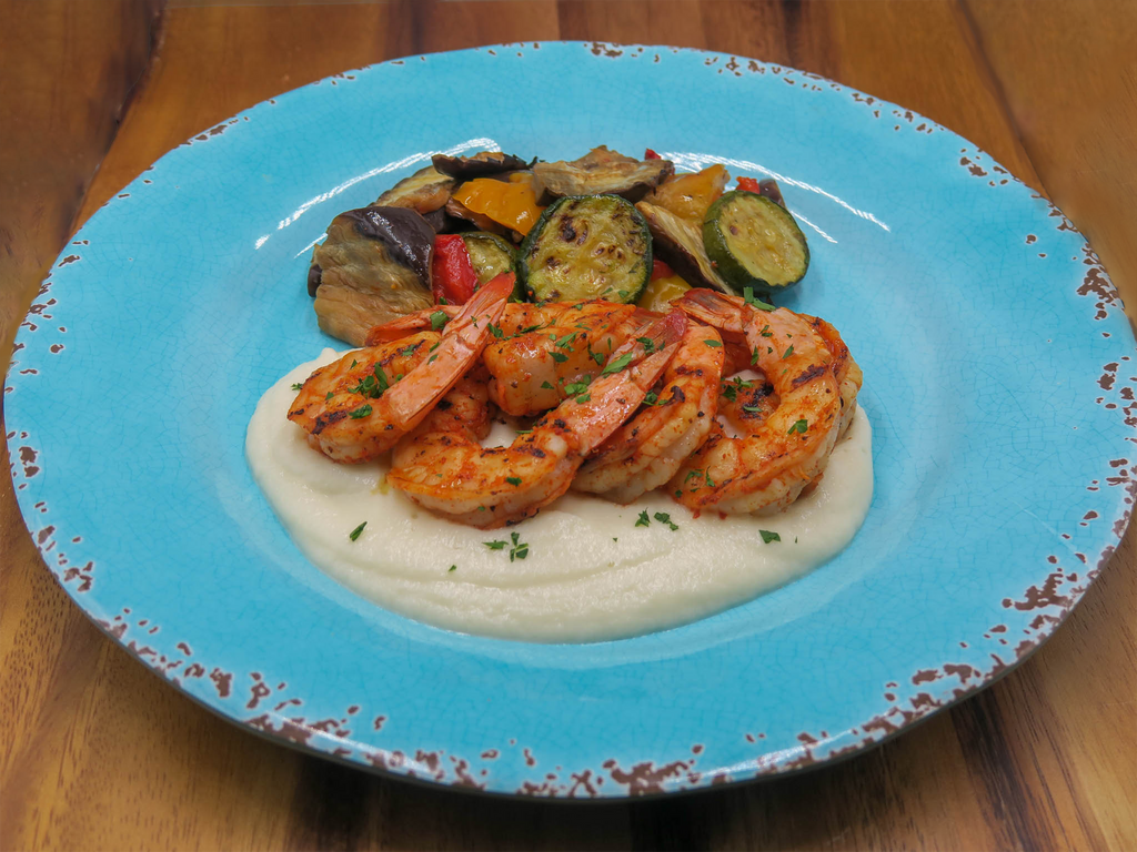 Grilled tail-on shrimp over cauliflower puree with a side of mixed grilled vegetables. Set on a blue plate on a brown cutting board.